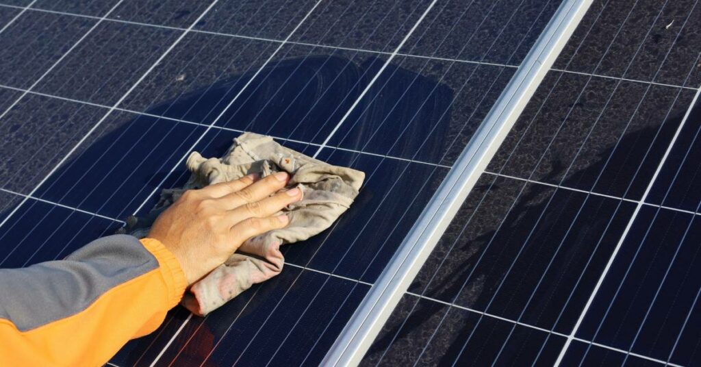 A woman's hand using a small, grey cloth to wipe off the dust, dirt, and grime from a set of solar panels.