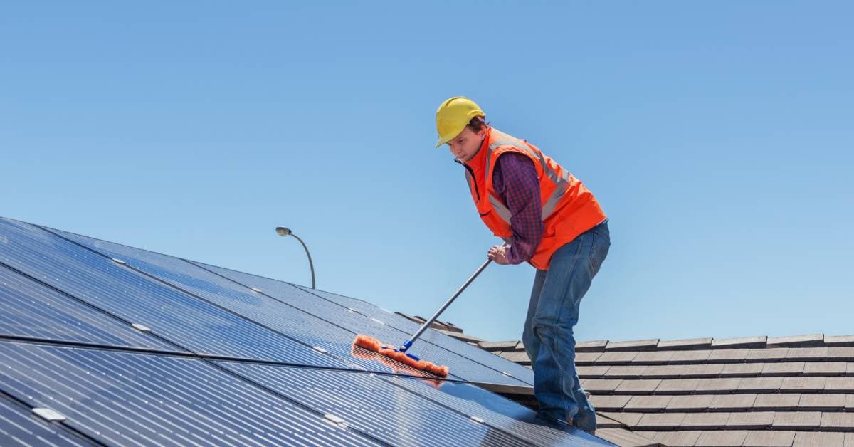 A woman in flannel, jeans, and orange high visibility gear using a dust mop to clean off the dust from rooftop solar panels.