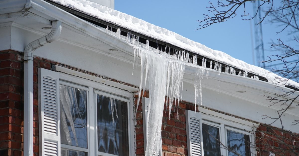 A red brick house with a snowy roof and large, long icicles pouring out of the broken gutter.