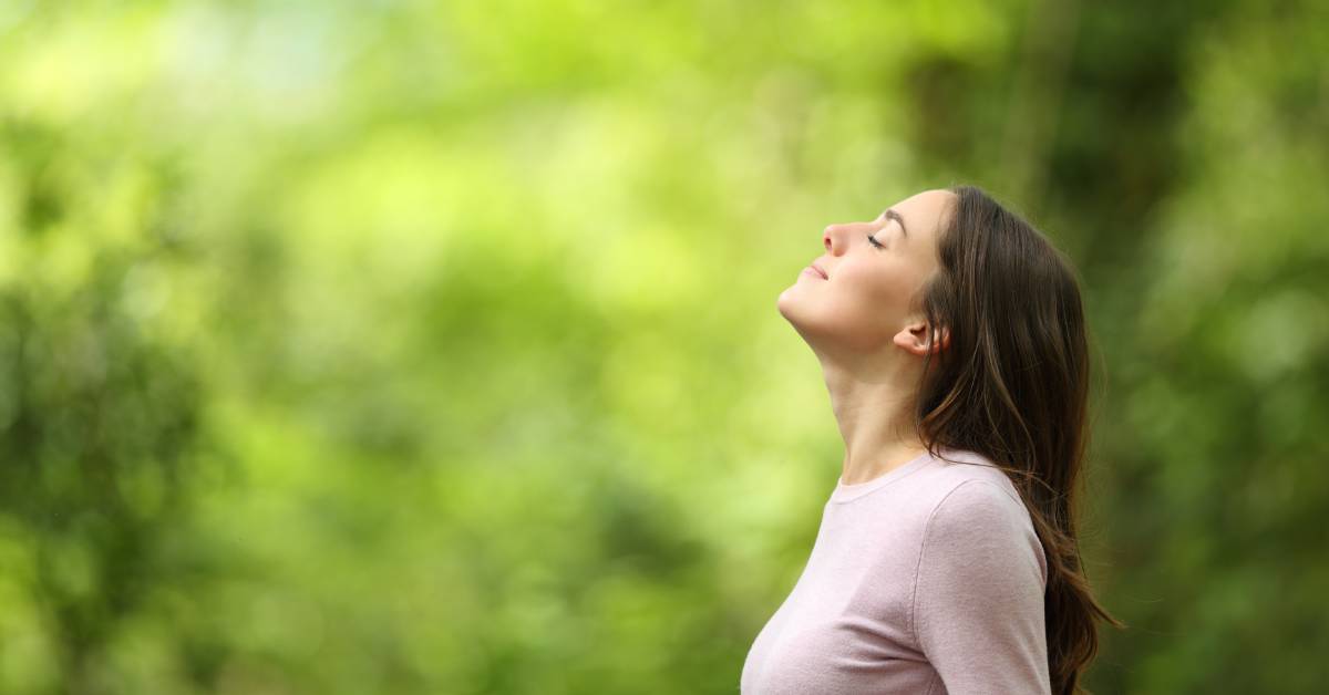 A woman is smiling and tilting her head up toward the sky. The background is blurred out, green, lush bushes and trees.