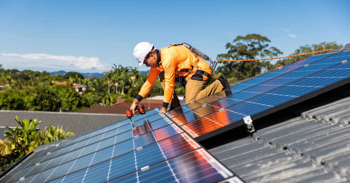 A worker in safety gear and an orange long-sleeved shirt is installing solar panels with a drill onto a rooftop.