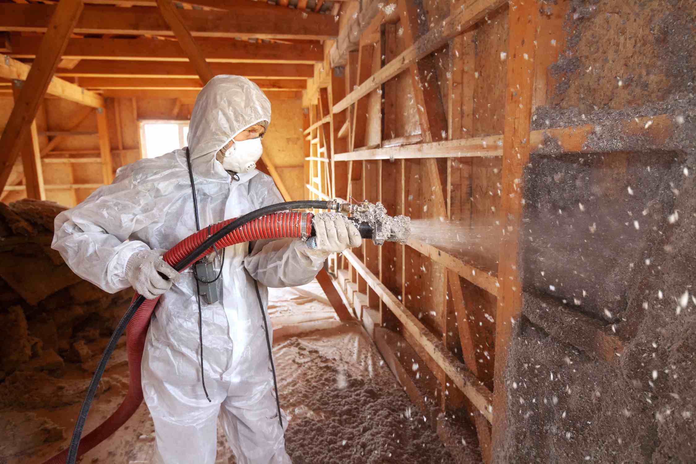 A worker wearing protective gear, using a large hose to spray cellulose insulation onto an attic wall.