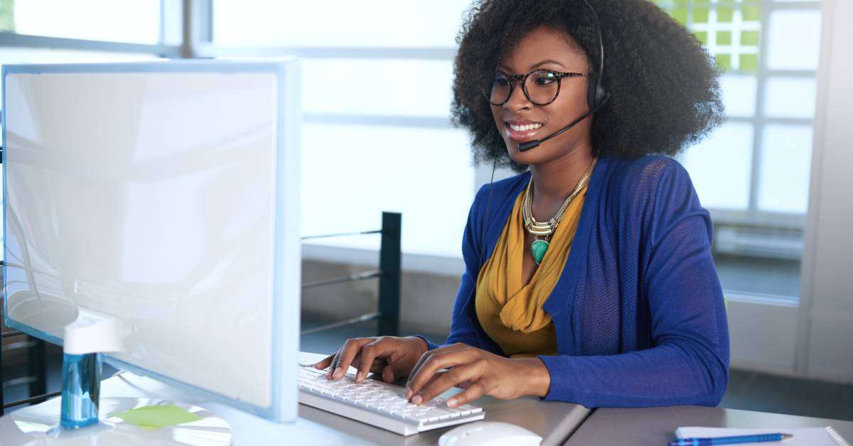 A portrait of a smiling customer service representative working at her computer desk while wearing a headset.