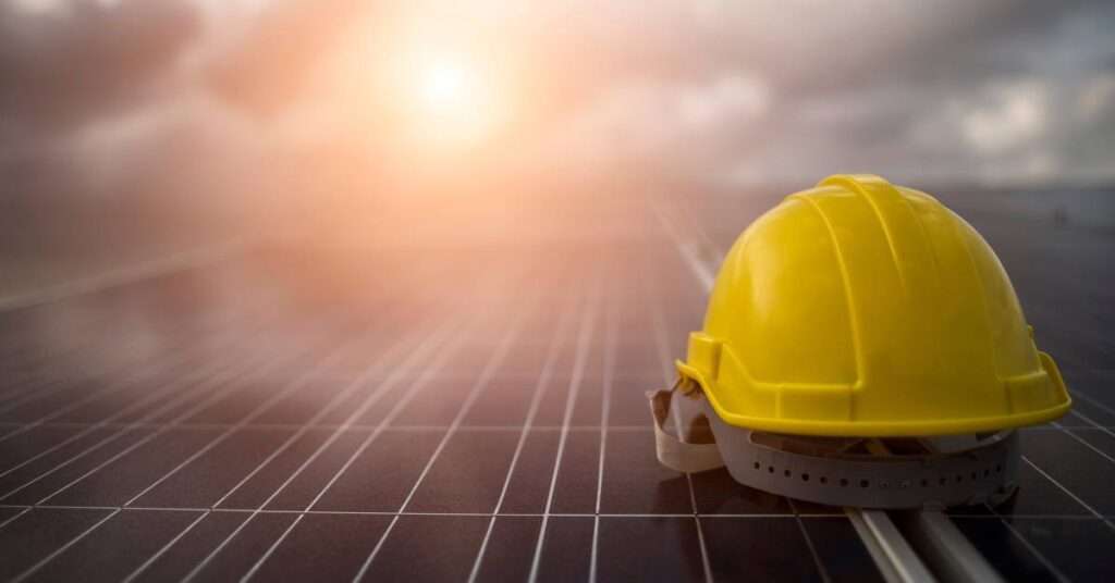 A yellow hard hat sitting on top of a set of solar panels with a grey sky and a bright sun in the background.