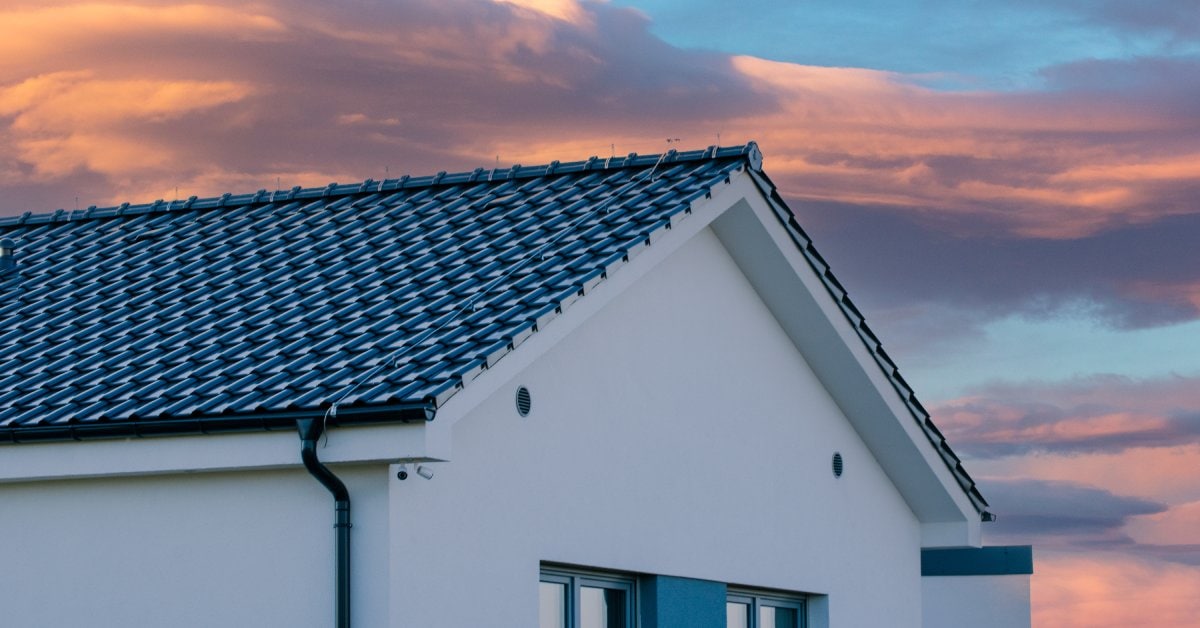 A white, modern home with black, sleek solar tiles over the original roof tiles. The sun sets in a pink sky in the background.