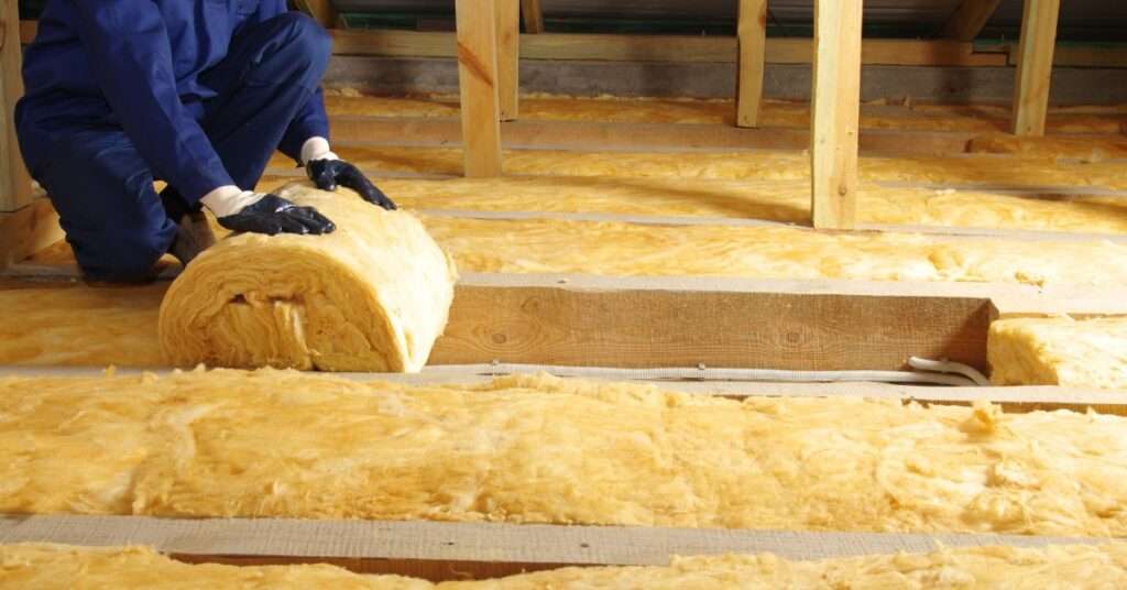 An unidentifiable worker in blue gear and protective gloves rolling yellow thermal insulation in an attic.