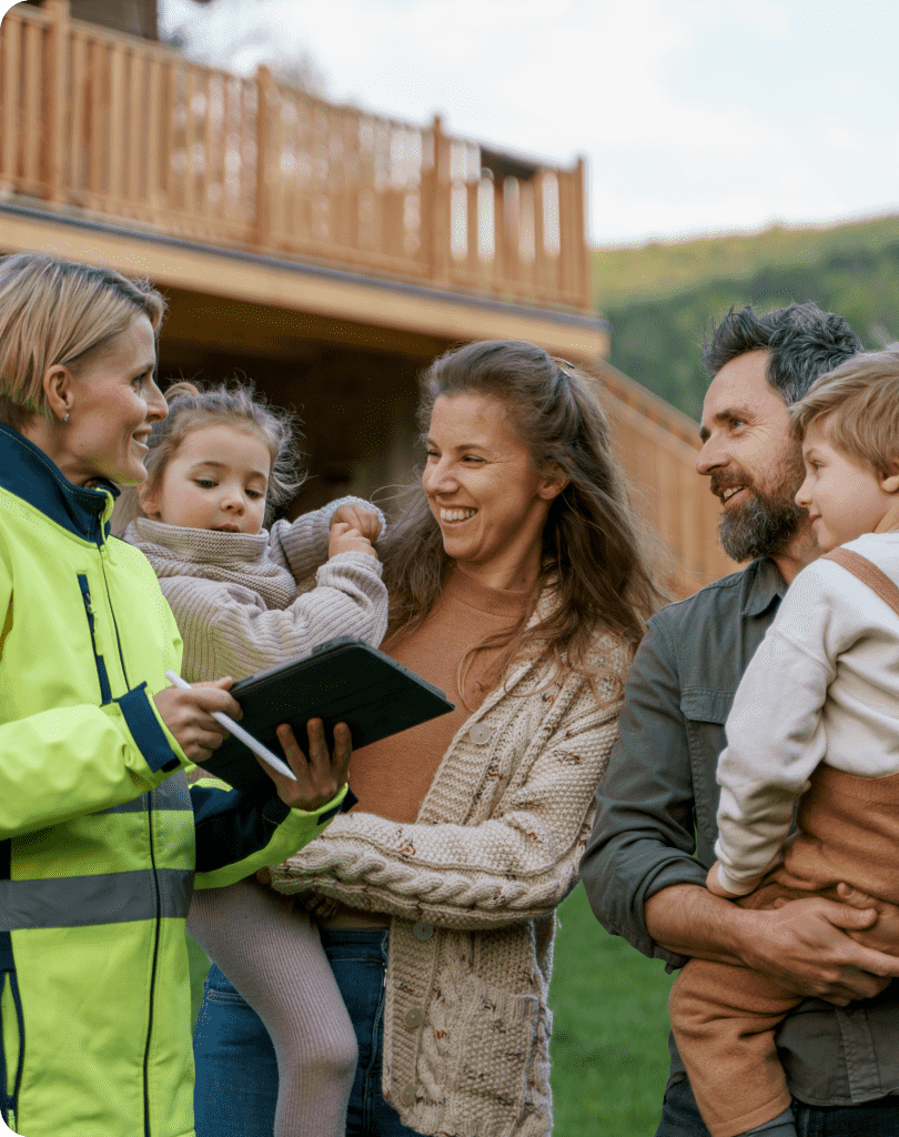 A woman in a high-visibility jacket holds a tablet and speaks with a smiling family of four, including two young children, outside a wooden house with a deck, discussing their new Greenlink installation.