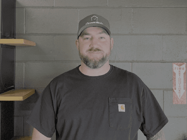 A bearded man in a black Carhartt t-shirt and gray cap stands by a gray brick wall near a fire extinguisher sign and empty wooden shelves, ready to offer solutions.
