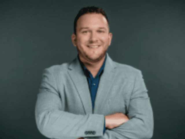 A man wearing a light gray blazer over a dark shirt stands with arms crossed and smiles confidently, ready to offer solutions, in front of a plain dark background.