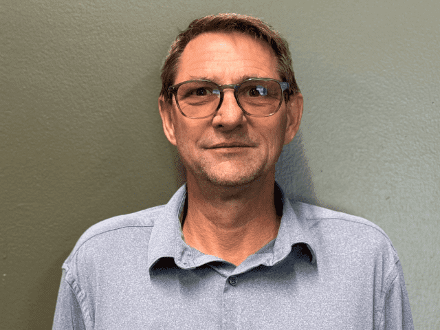 A man wearing glasses and a light blue collared shirt stands in front of a plain, light green wall, looking directly at the camera with a neutral expression, as if ready to discuss solutions.