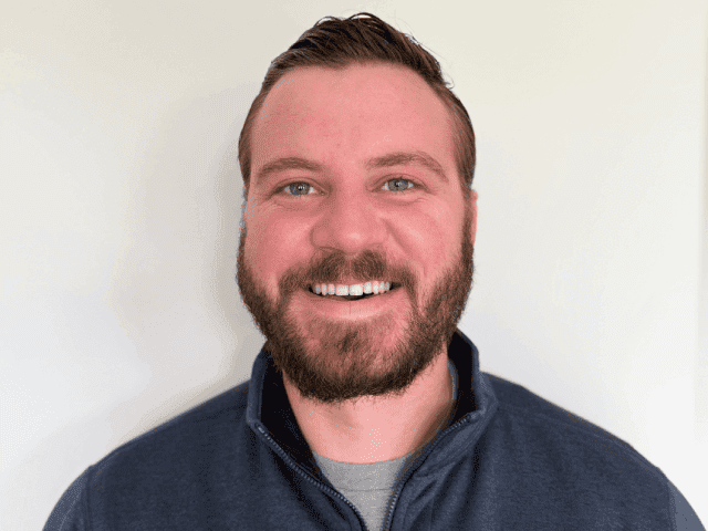 A man with short brown hair and a beard is smiling at the camera, ready to share solutions. He is wearing a dark zip-up jacket over a gray shirt, standing in front of a plain white background.
