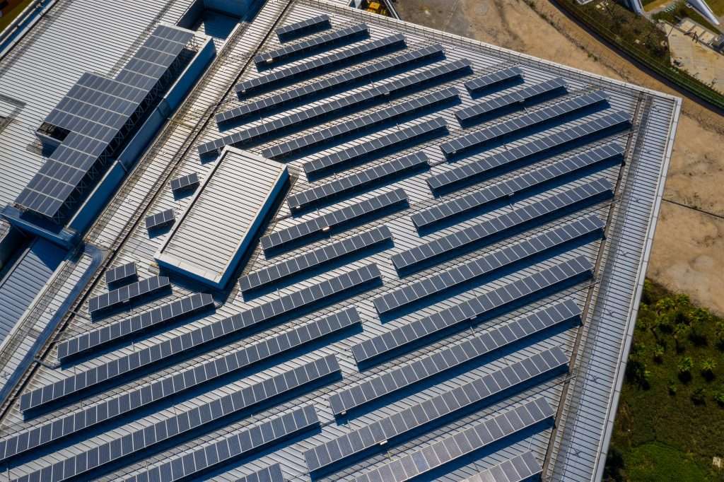 Aerial view of a large industrial building roof covered with multiple rows of solar panels; solar energy powers the site, with some greenery and bare ground visible at the edge of the structure.