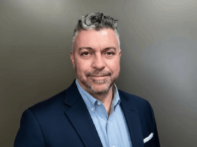 A man with short, curly gray hair and a trimmed beard wears a dark suit jacket over a light blue dress shirt, standing in front of a plain gray background, facing the camera and smiling slightly, ready to offer insightful solutions.