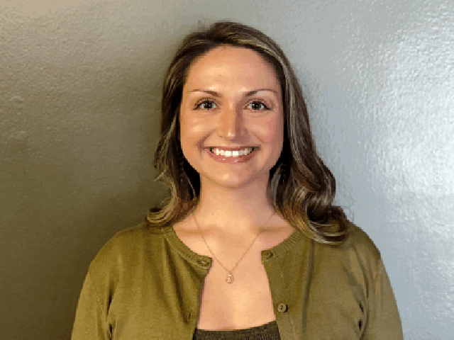 A woman with light brown hair, wearing a green cardigan and a necklace, stands in front of a plain, light gray background, smiling confidently at the camera, ready to share her solutions.