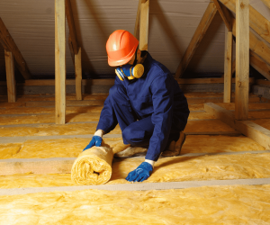 A person wearing a hard hat, protective mask, gloves, and coveralls is kneeling in an attic and installing fiberglass insulation between wooden beams. A person wearing a hard hat, protective mask, gloves, and coveralls is kneeling in an attic and installing fiberglass insulation between wooden beams.