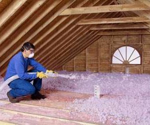 A person wearing safety glasses, a mask, gloves, and a blue shirt is using a hose to install GreenLink loose-fill insulation in an unfinished attic with exposed wooden beams and a small window. A person wearing safety glasses, a mask, gloves, and a blue shirt is using a hose to install GreenLink loose-fill insulation in an unfinished attic with exposed wooden beams and a small window.